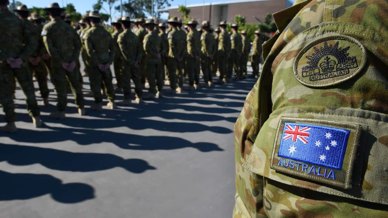 The Australian flag on the sleeve of a soldier's uniform in the foreground. Soldiers are standing in rows in the background.