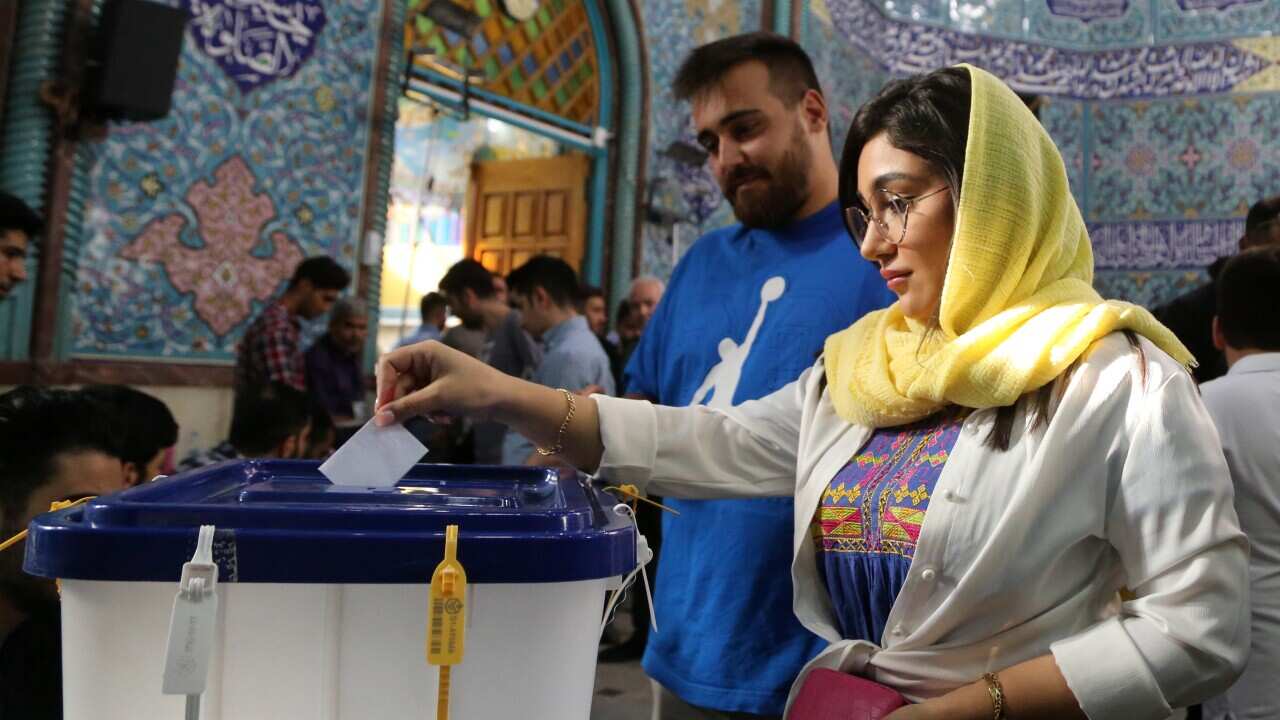 A woman putting a piece of paper into a ballot box at a polling station. A man in a blue shirt is behind her.