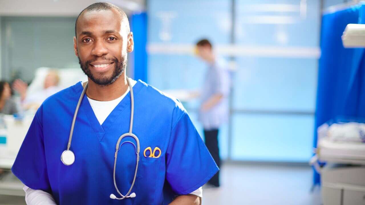 a mid adult doctor looks to camera on the hospital ward ,with a young male nurse , patient , and relative in the background.