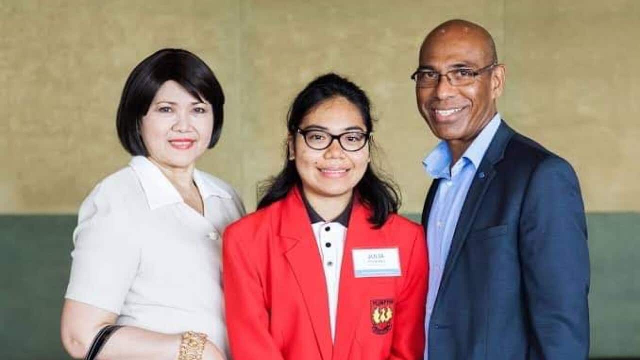 A young woman with glasses wearing a red blazer stands between an older woman and man (her mother and father). The mother is wearing a white shirt, the father a light blue shirt and dark blue suit jacket. 