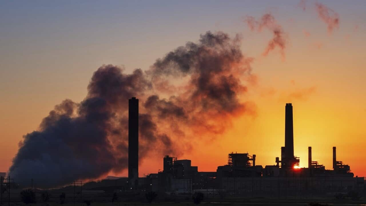 A cloud of smoke billows in front of a sunset and industrial site.