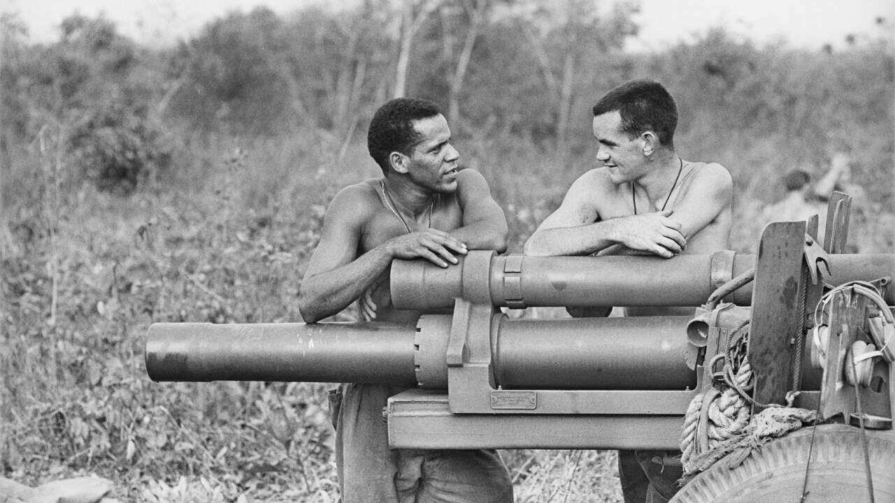 Two shirtless men, one black and one white, casually speak while leaning on artillery guns in a Vietnamese field. 