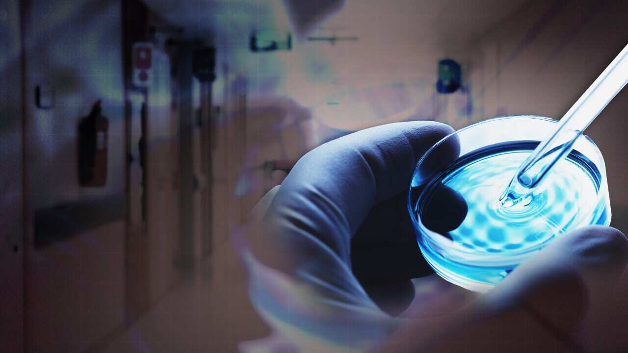 Person wearing gloves holding a small pot of liquid, with a hospital corridor in the background. 