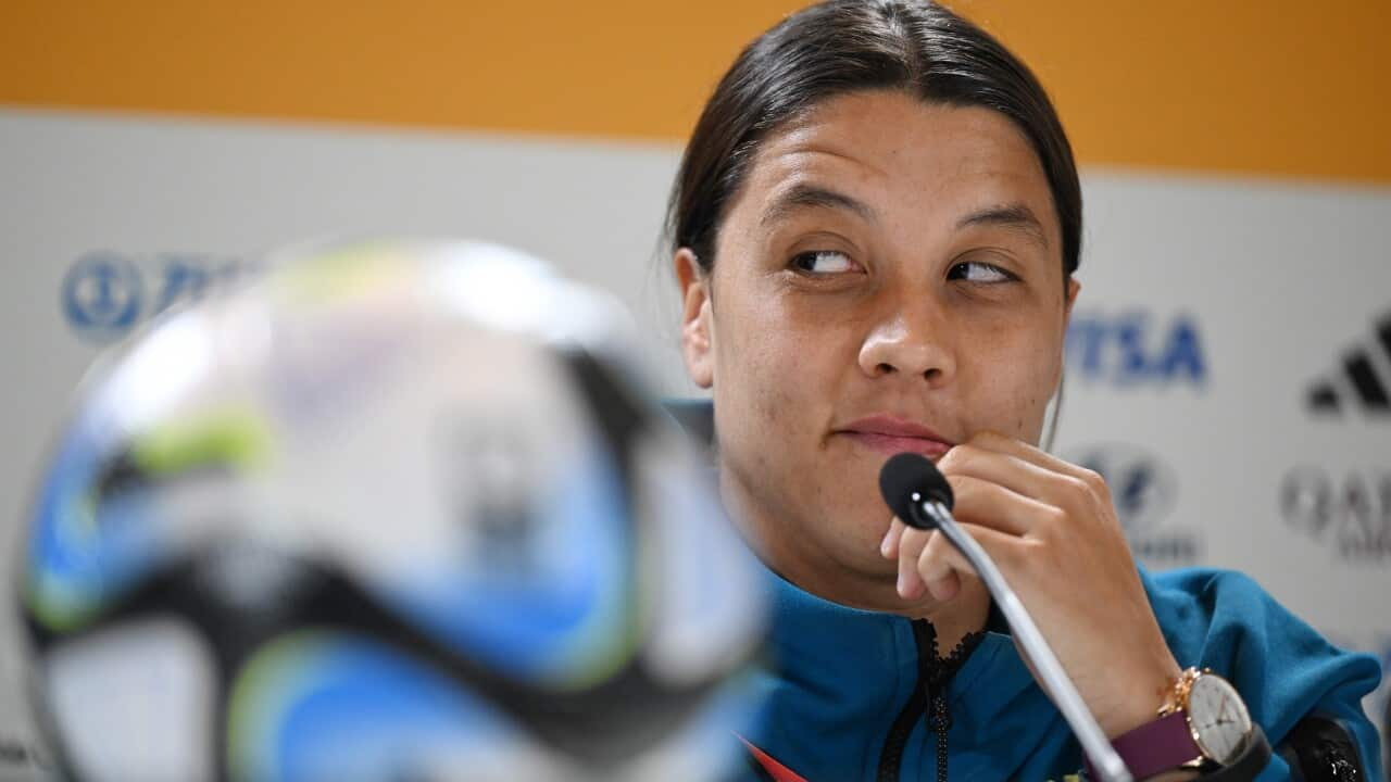 Sam Kerr, wearing a tracksuit, sitting behind a microphone at a press conference. There is a football on the table in front of her.