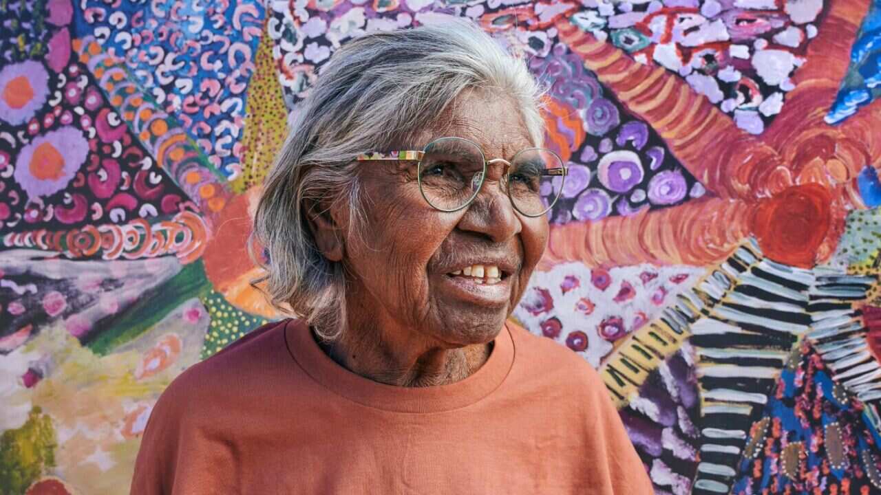 An older woman with white hair standing in front of a vibrant artwork, wearing glasses that mirror the work's design. 