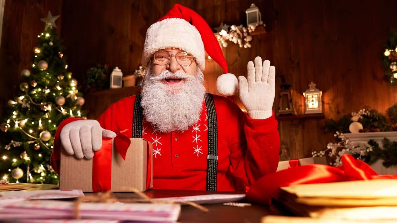 Santa Claus at a desk piled with envelopes and packages, smiling and waving at the camera