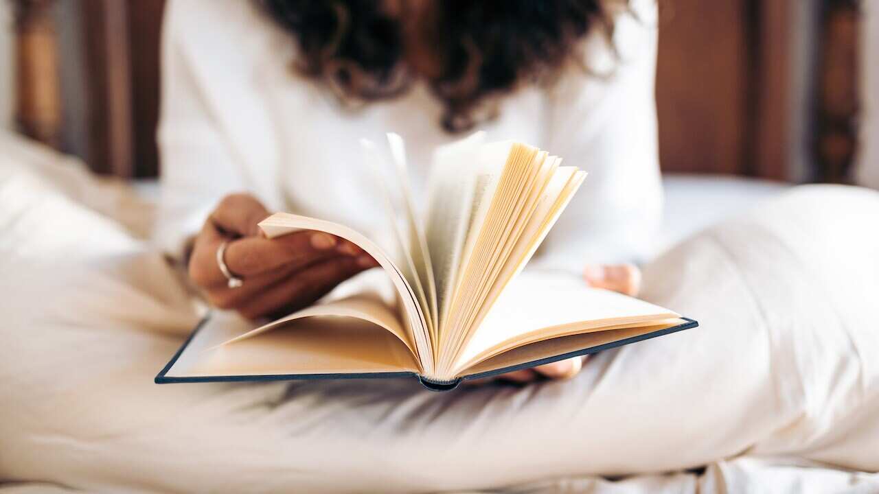 Young woman turning the pages of a book while reading in bed covered with the duvet