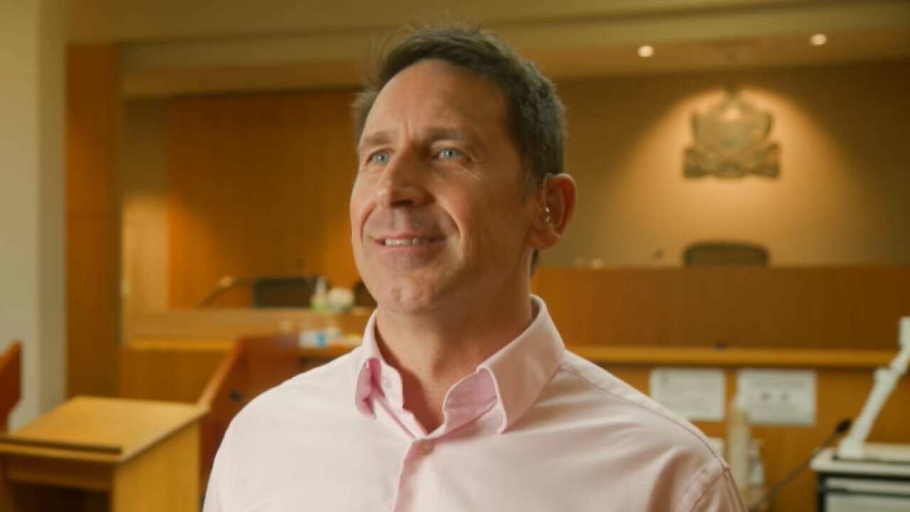 Dressed in a light pink business shirt, a man uses sign language in a courtroom to respond to questions from SBS News during an interview. 