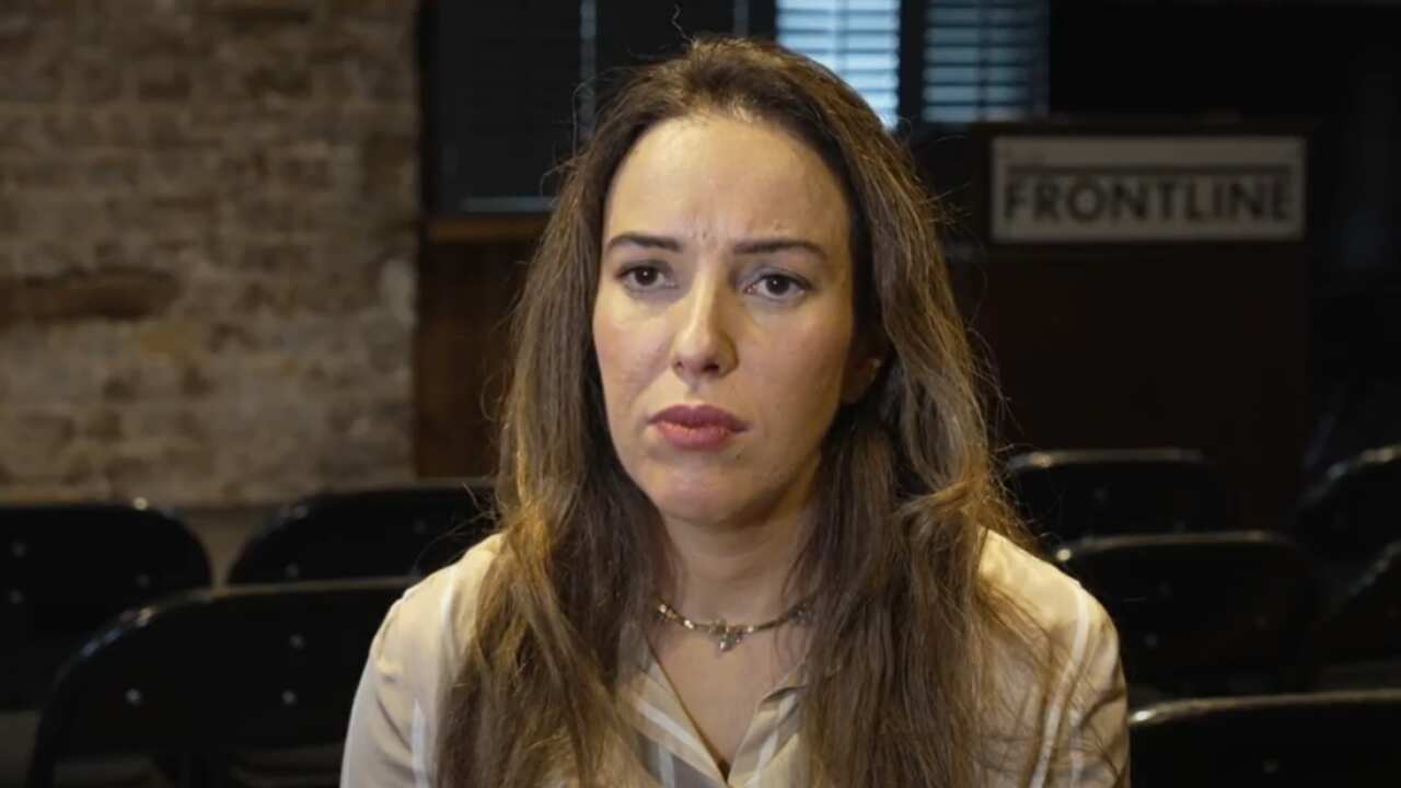 A woman is seated in front of a camera speaking to a journalist.