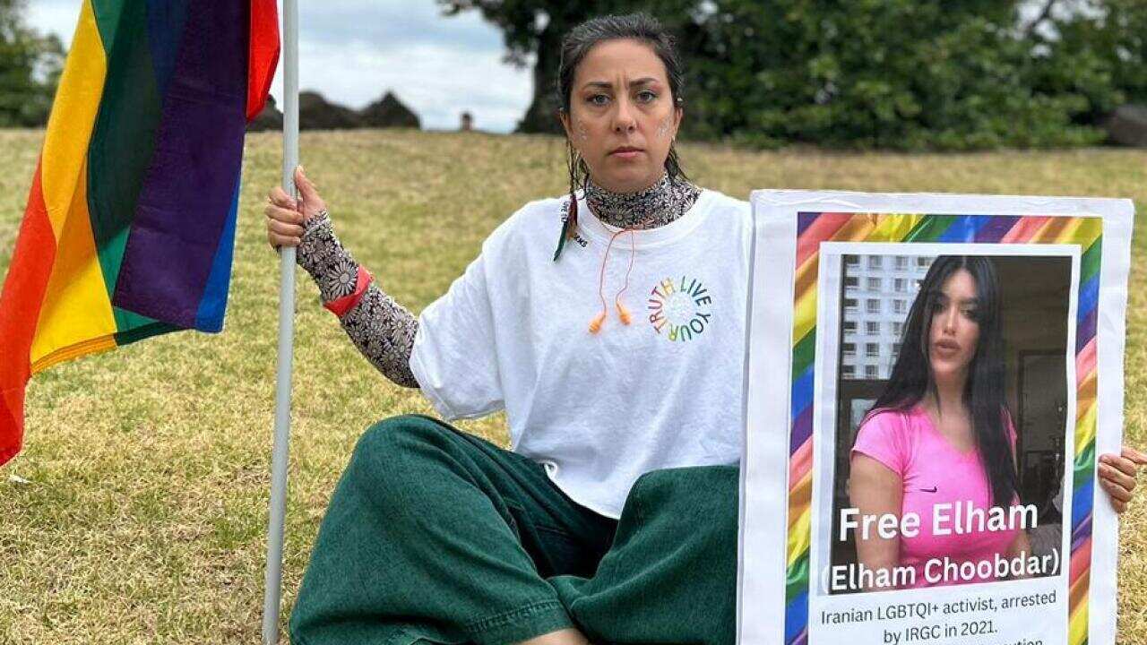 A woman sits on the grass carrying a poster and a pride flag. 