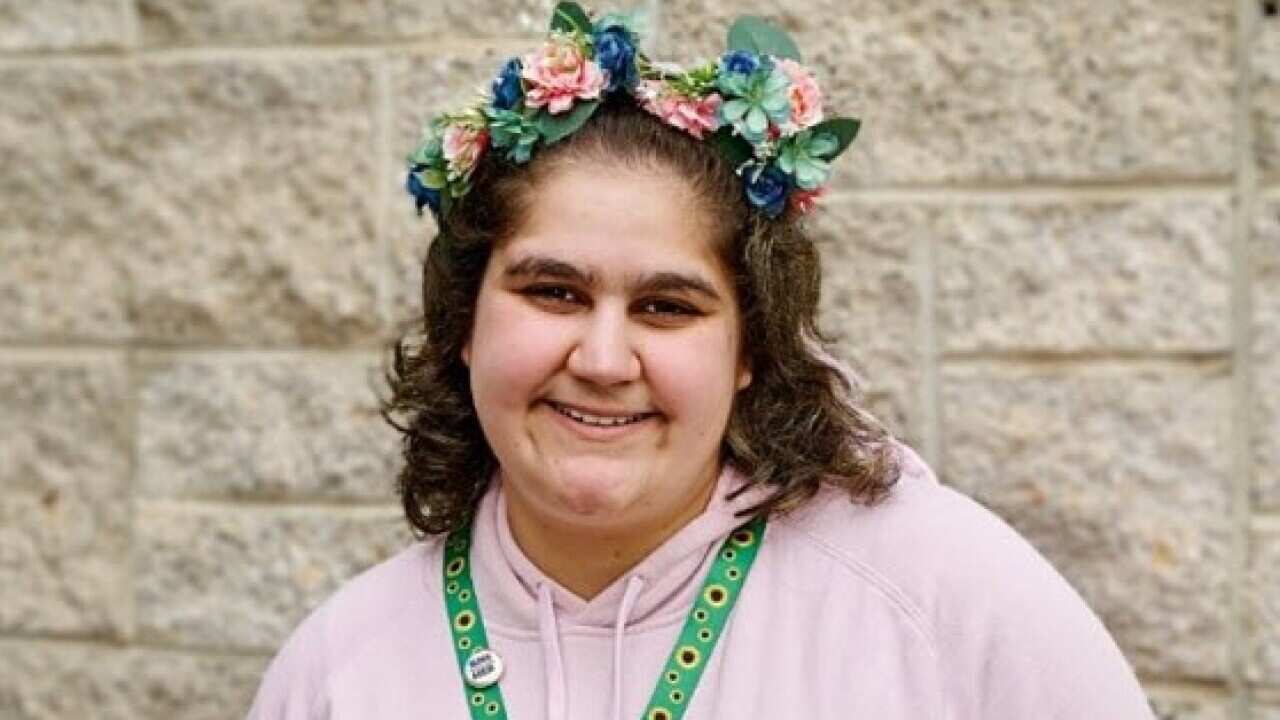 A young woman in a pink hoodie with a flower crown around her head and a sunflower lanyard around her neck