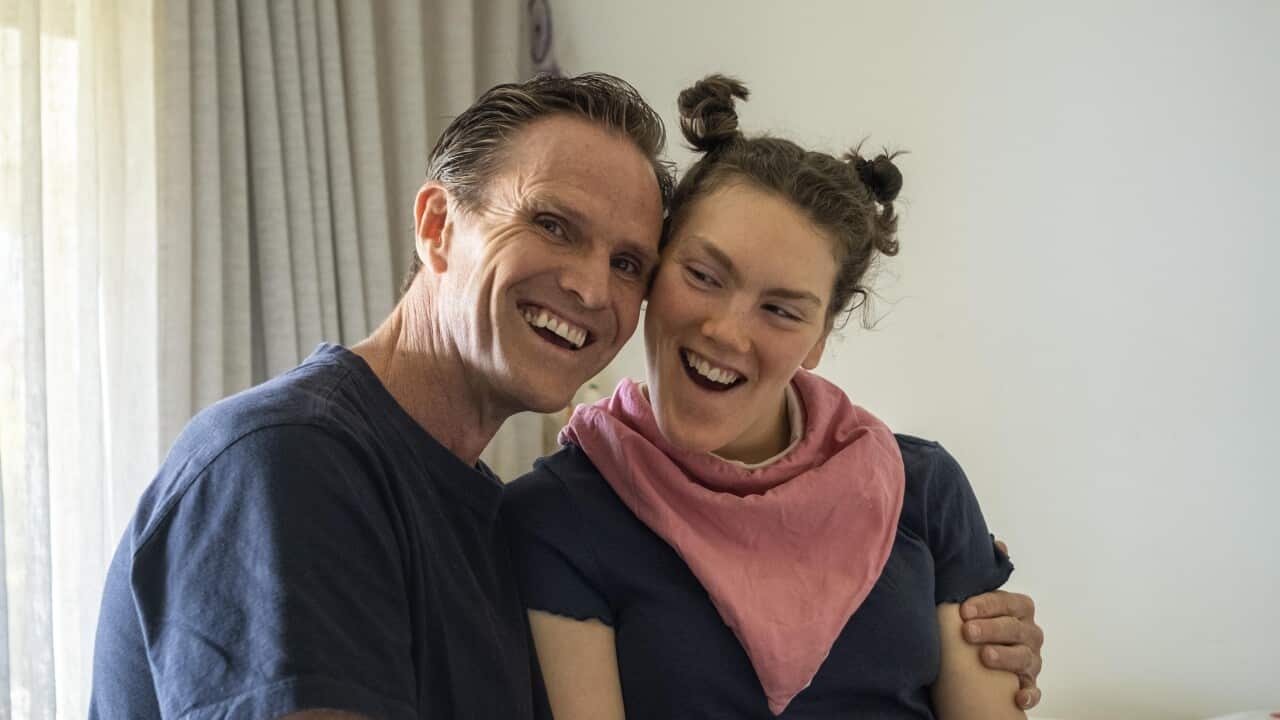 Image of a man wearing a blue tshirt and khaki shorts with his arm wrapped around his daughter who has her hair in two buns on top of her head, a pink scarf around her neck and a navy tshirt. They are both smiling widely.