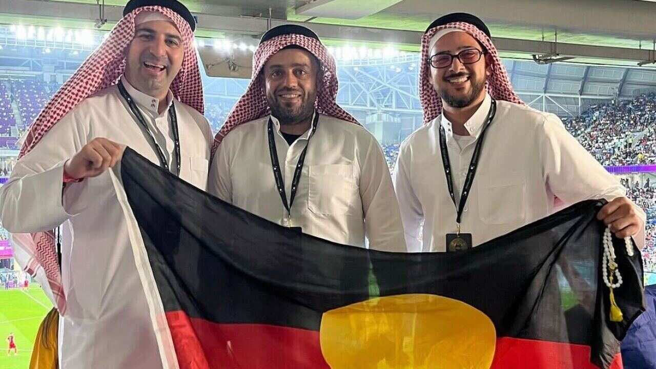 Three men in thobes carry an Aboriginal flag. 