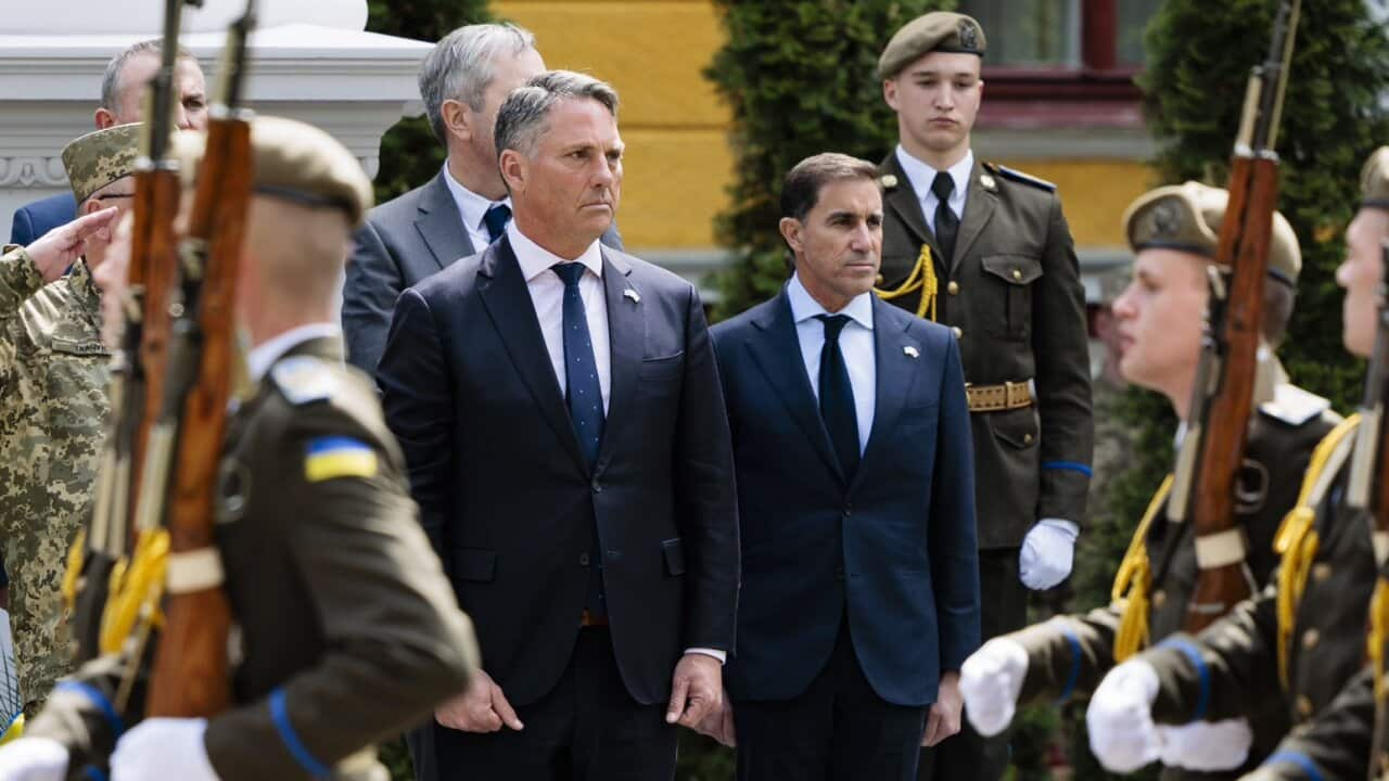 Two men in suits stand while Ukrainian soldiers holding rifles walk past them