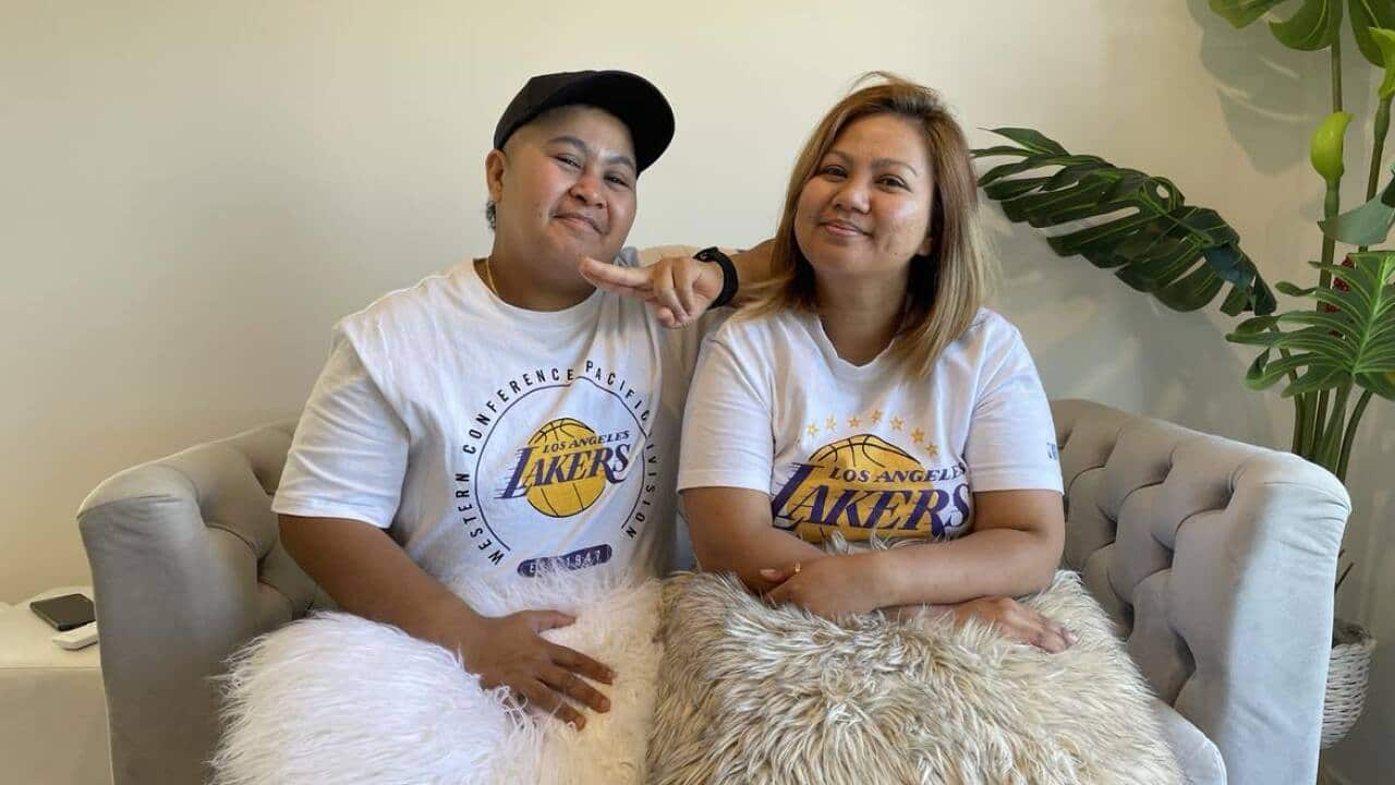 Two people in white Lakers T-shirts with fluffy pillows on their laps sit on a couch smiling. 