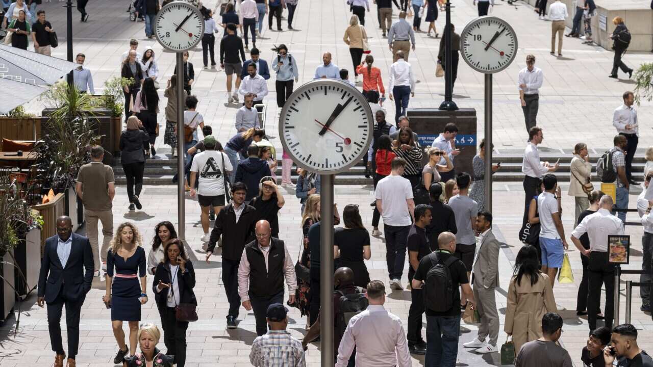 A crowd of people walk past several analog clocks raised on metal poles at the canary wharf in London