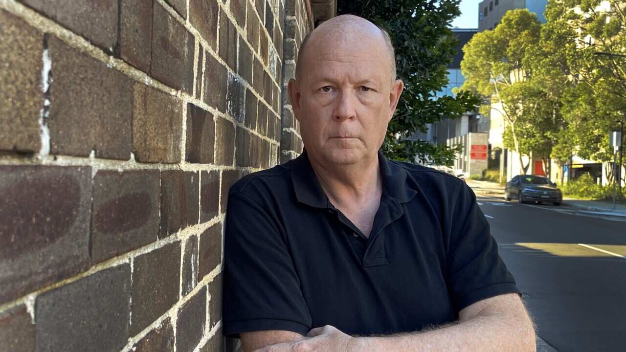 A man with a serious expression leans against a wall.