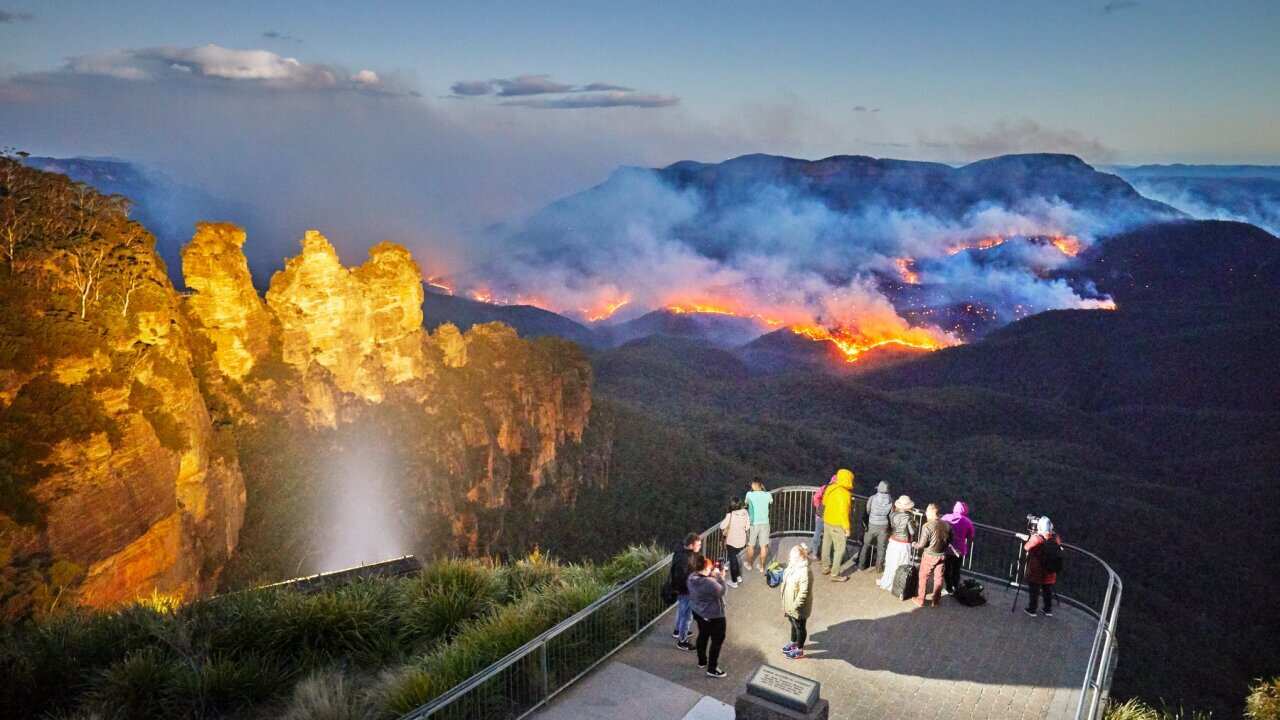 Three Sisters floodlit at dusk, Queen Elizabeth Lookout, viewing platform with tourists watching and photographing fire, bushfire in Jamison Valley, Blue Mountains National Park, Australia