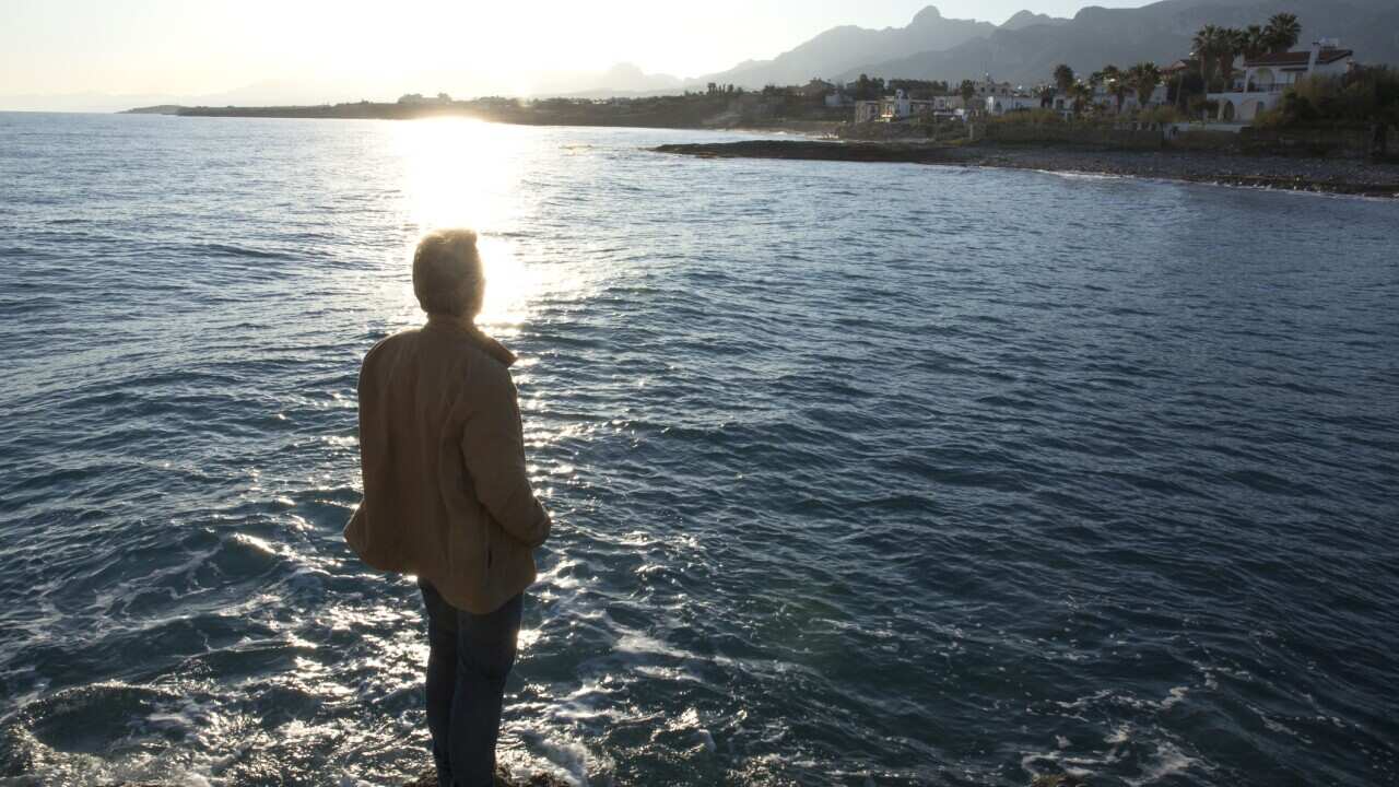 Man looks across sea & distant village,from shore