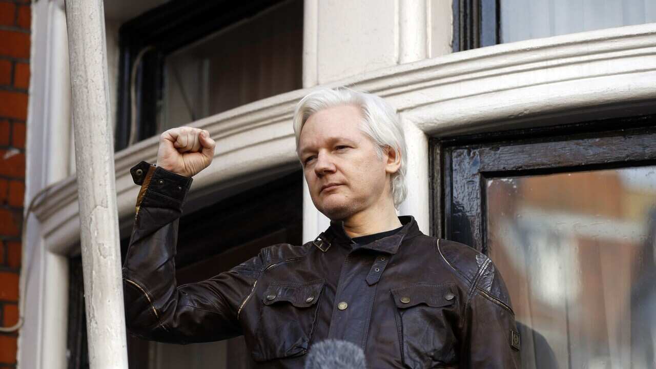 Julian Assange stands on a balcony with curved windows behind him. He is wearing a black jacket and holding up his right fist.