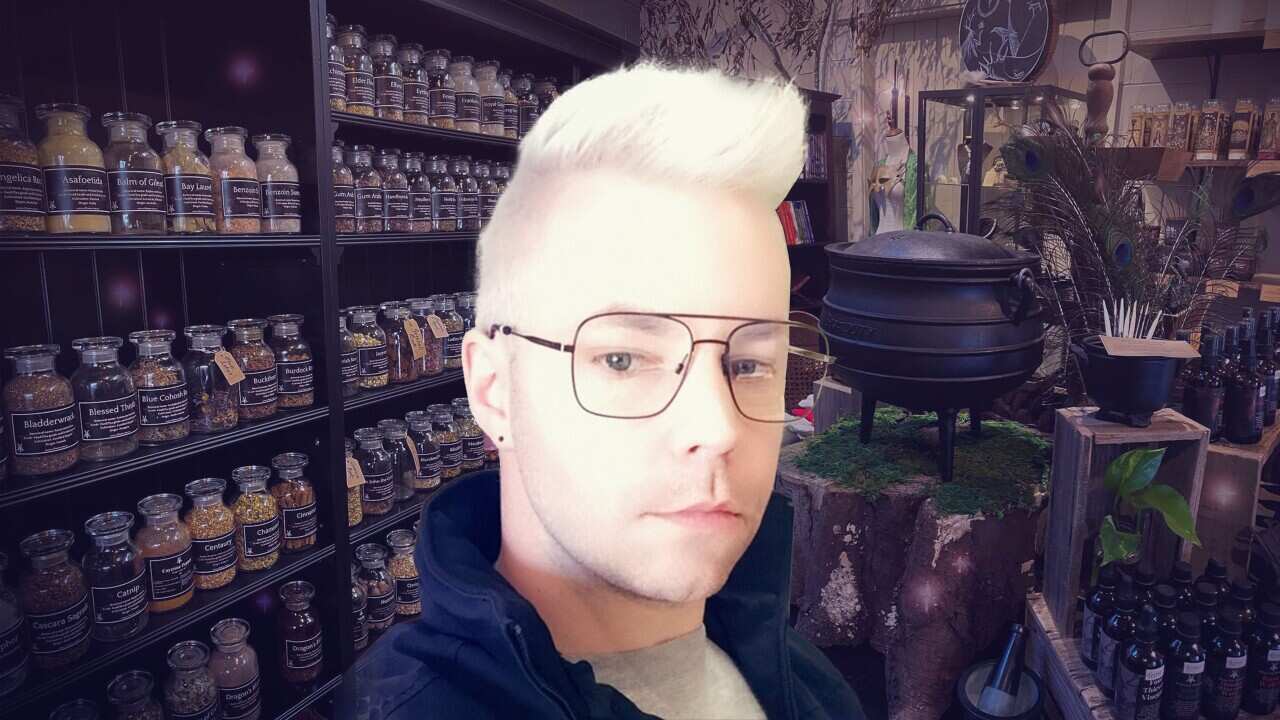 A man with bleached hair and glasses stares into the camera. Behind him are a cauldron, rows of black bottles and dark wooden shelves filled with glass jars of herbs.