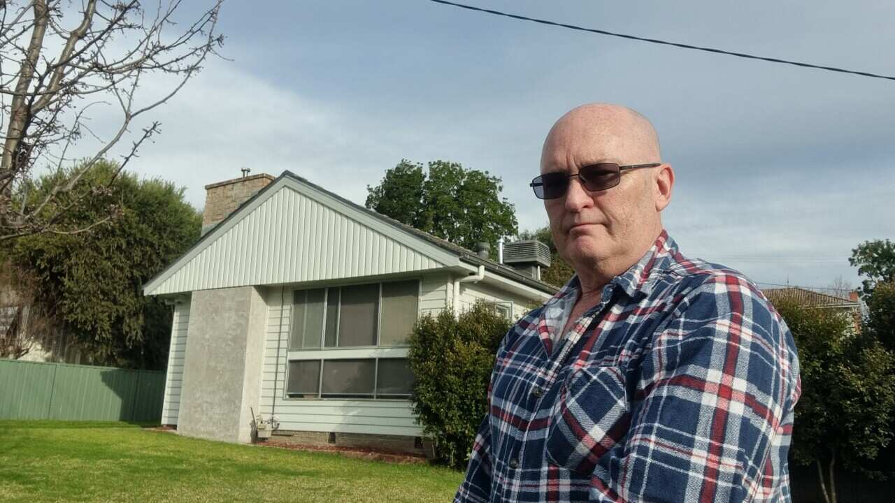 A man in a checked shirt and sunglasses stands in front of modest home.