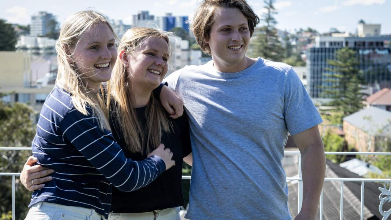 image of a blonde woman in a striped t-shirt hugging a blonde woman wearing a black t-shirt next to their brother who is wearing a grey t-shirt and board shorts. All standing outside on a balcony in the sun,