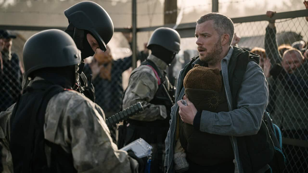 A man stands in front of a chain fence, talking to three people in soldier's uniforms. 