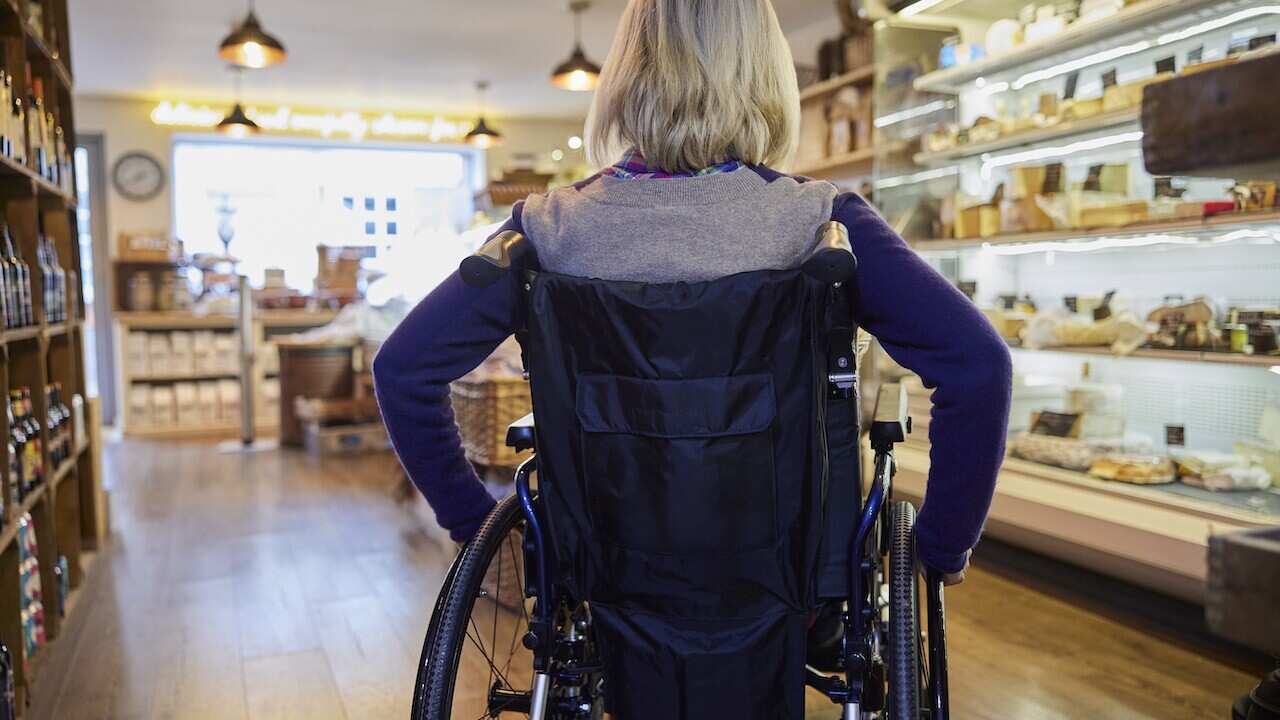 Rear View Of Woman In Wheelchair Shopping For Food In Delicatessen