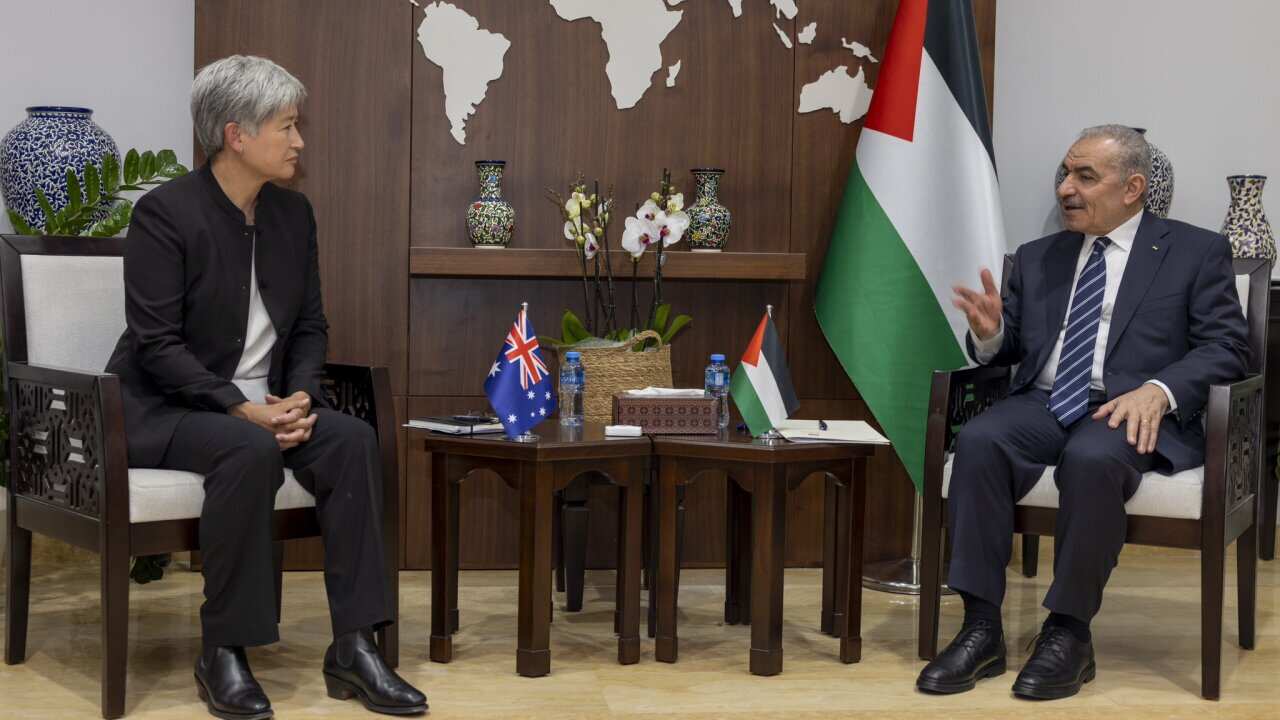 A man and a woman, both wearing formal attire, sit facing each other. A map of the world and Palestinian flag are visible behind them. 