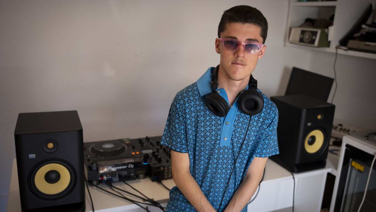 A teenage boy wearing pink sunglasses, a blue patterned polo shirt, and with headphones around his neck stands in front of some DJing equipment.