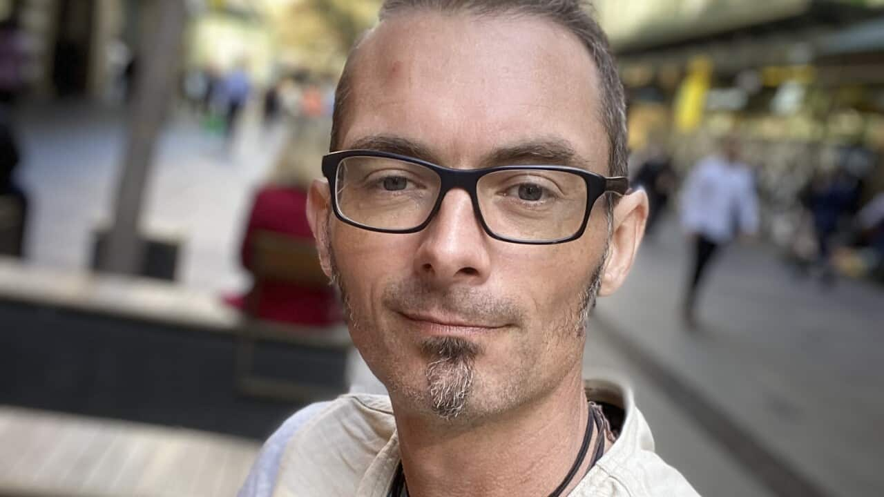 A man with glasses wearing a white T-shirt stands in the street and takes a selfie.