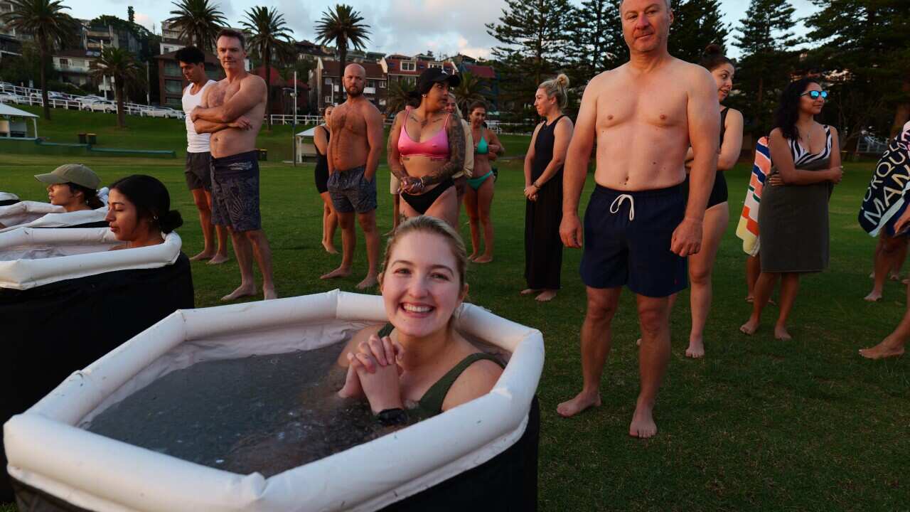 A woman sits in an ice bath and smiles, while around her others are sitting in baths or waiting to get in.