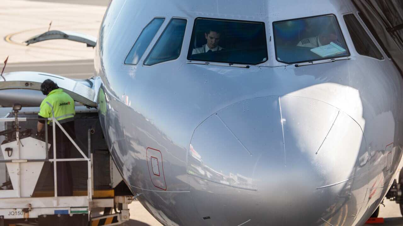 Pilots sit in the cockpit of an aircraft at an airport with an airport crew in green vest working on the left.