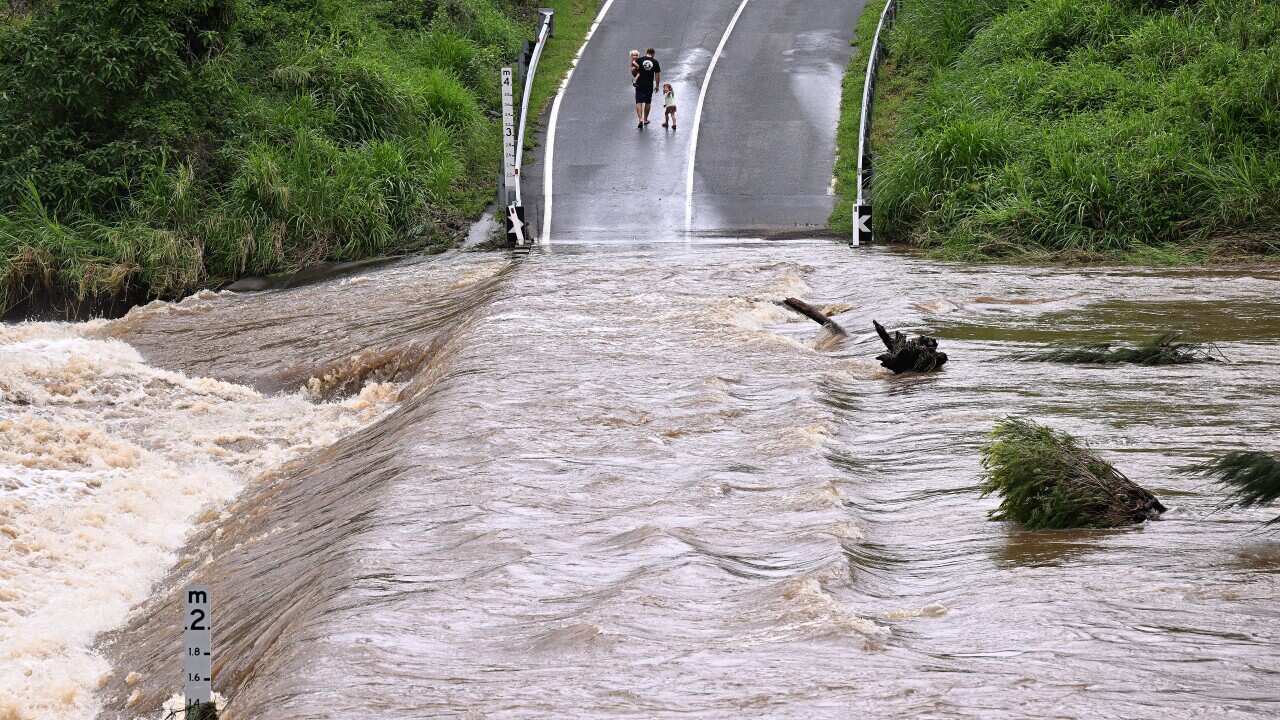 A man and his two children in the distance walking along a pathway, with rushing floodwaters in the distance.