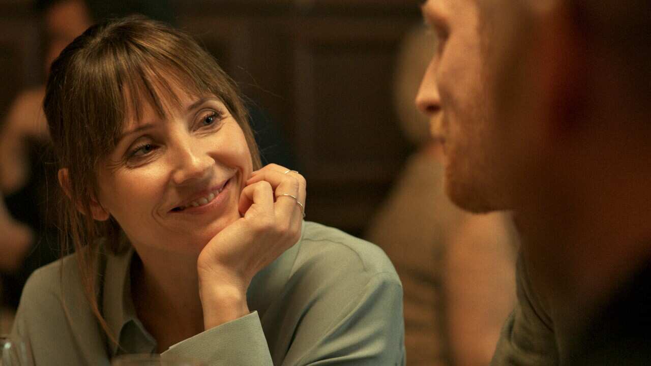 A smiling woman leans with her hand under her chip, smiling at a man who is partly seen, in profile. 