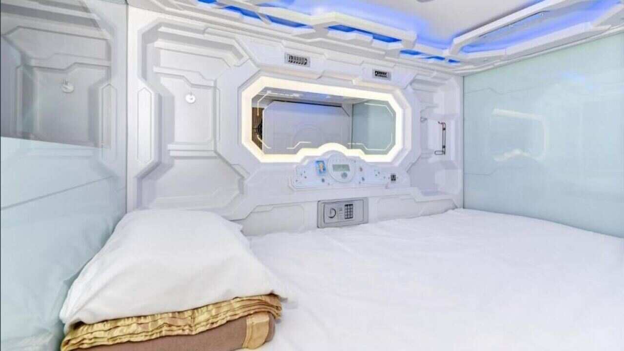 A small, futuristic-looking room with a mattress covering the entire space, a pillow and a window to a hallway.