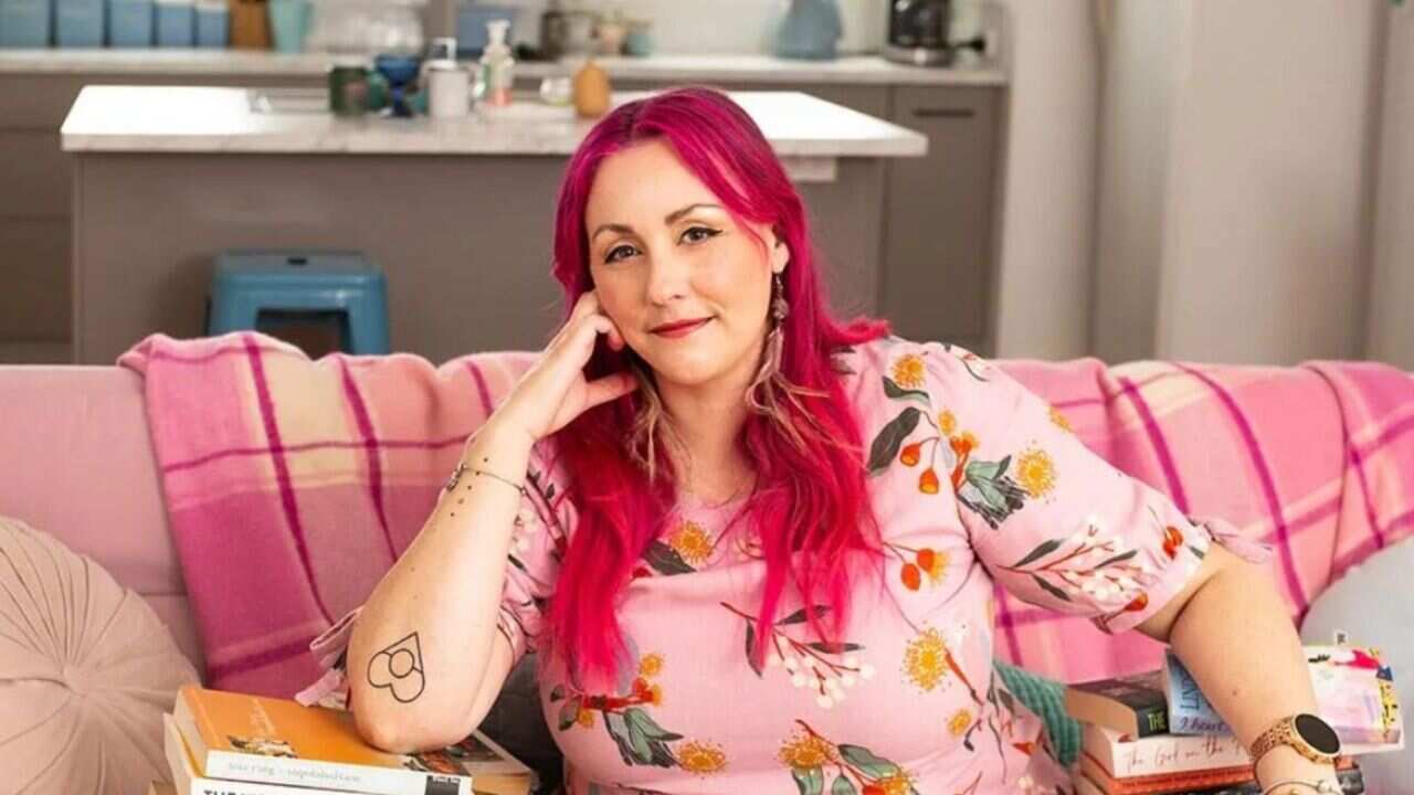 Bigambul and Wakka Wakka author Melanie Saward has bright pink hair and dark brown eyes. She sits on a pink lounge surrounded by stacks of books, looking into the camera.