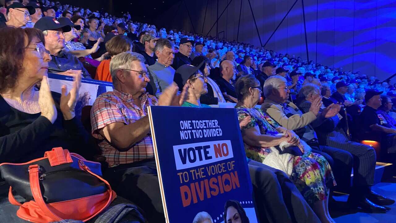 A crowd of people sit in an auditorium, one with a 'Vote No' placard