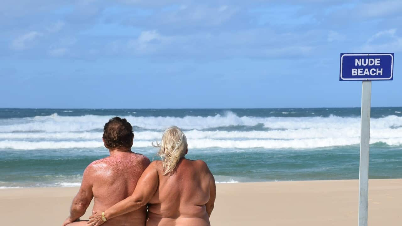 Photo taken from behind of a man and woman on a beach with no shirts on, and a sign saying 'nude beach'. 