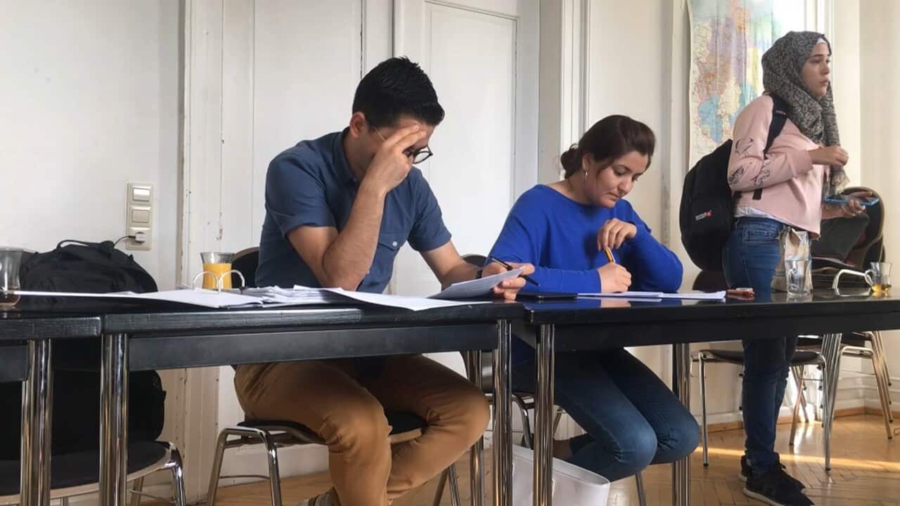 A man and a women sitting at a desk. A woman in a headscarf is standing.