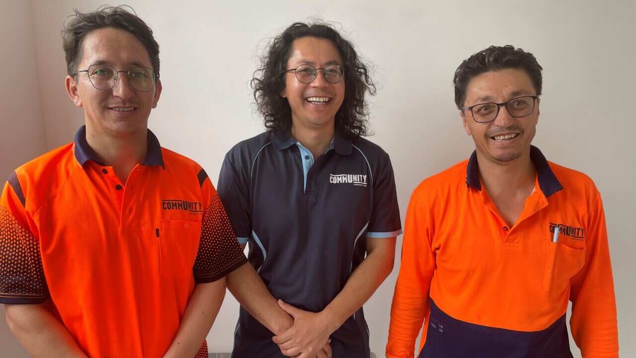 Three men standing in a row inside. The ones on the left and right are wearing orange hi-vis shirts and the one in the middle is wearing a dark blue polo shirt.