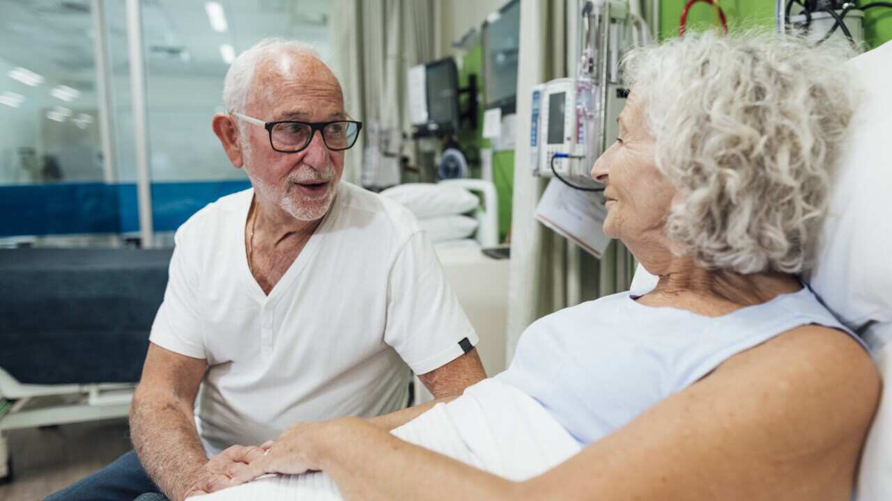 An older woman in a hospital bed speaking to an older man.