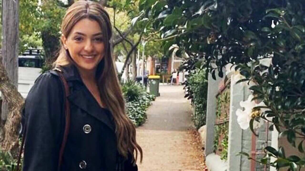 Young woman with long brown hair smiling at camera in leafy street.