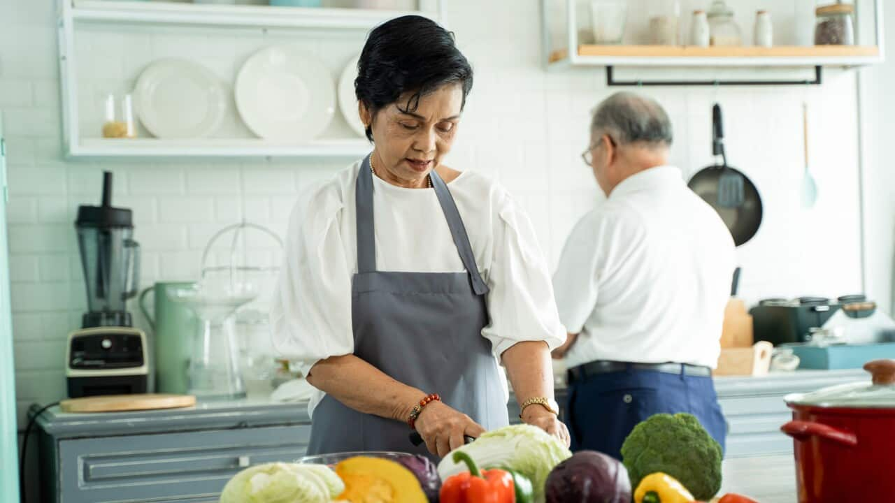 Elderly Asian woman cooking and cutting ingredients on table while preparing lunch with husband in cozy kitchen at home. Senior Asian couple cooking together