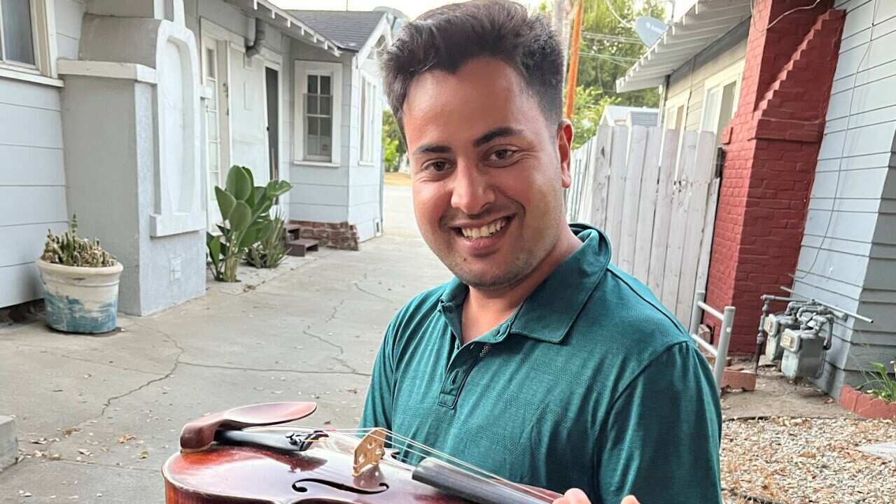 A smiling man standing in a laneway, taking a violin out of its case.  